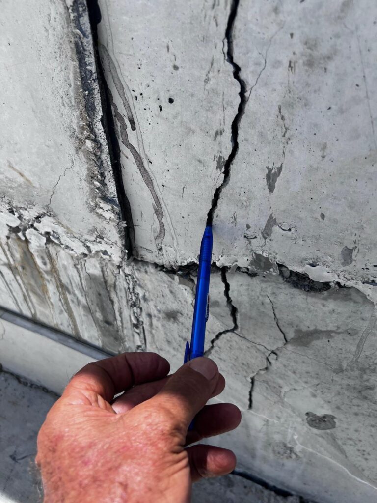 A hand pointing a blue ink pen at cracks along a concrete wall.